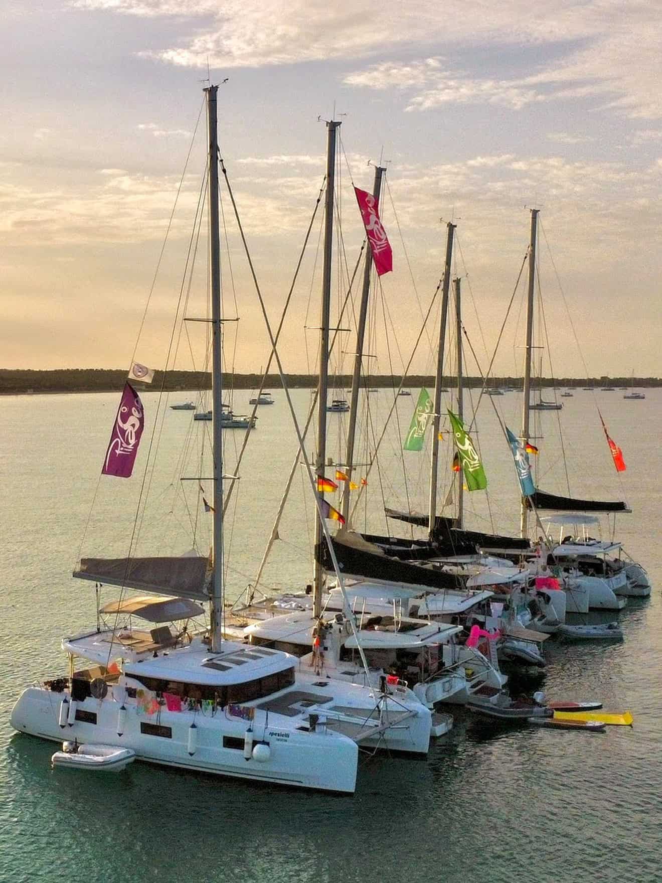 Colorful sailing yachts moored at sunset on the calm sea, with vibrant flags flying from their masts, creating a lively and picturesque maritime scene.