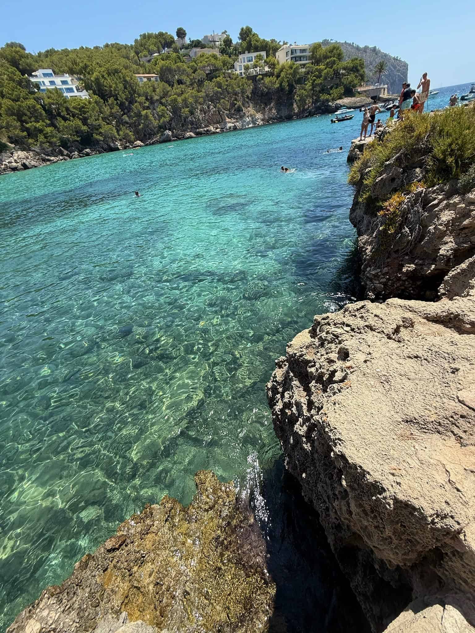 Secluded Mediterranean cove with clear turquoise waters, rocky shoreline, and lush green hillside with modern white villas, perfect for relaxing, swimming, and enjoying scenic coastal views.