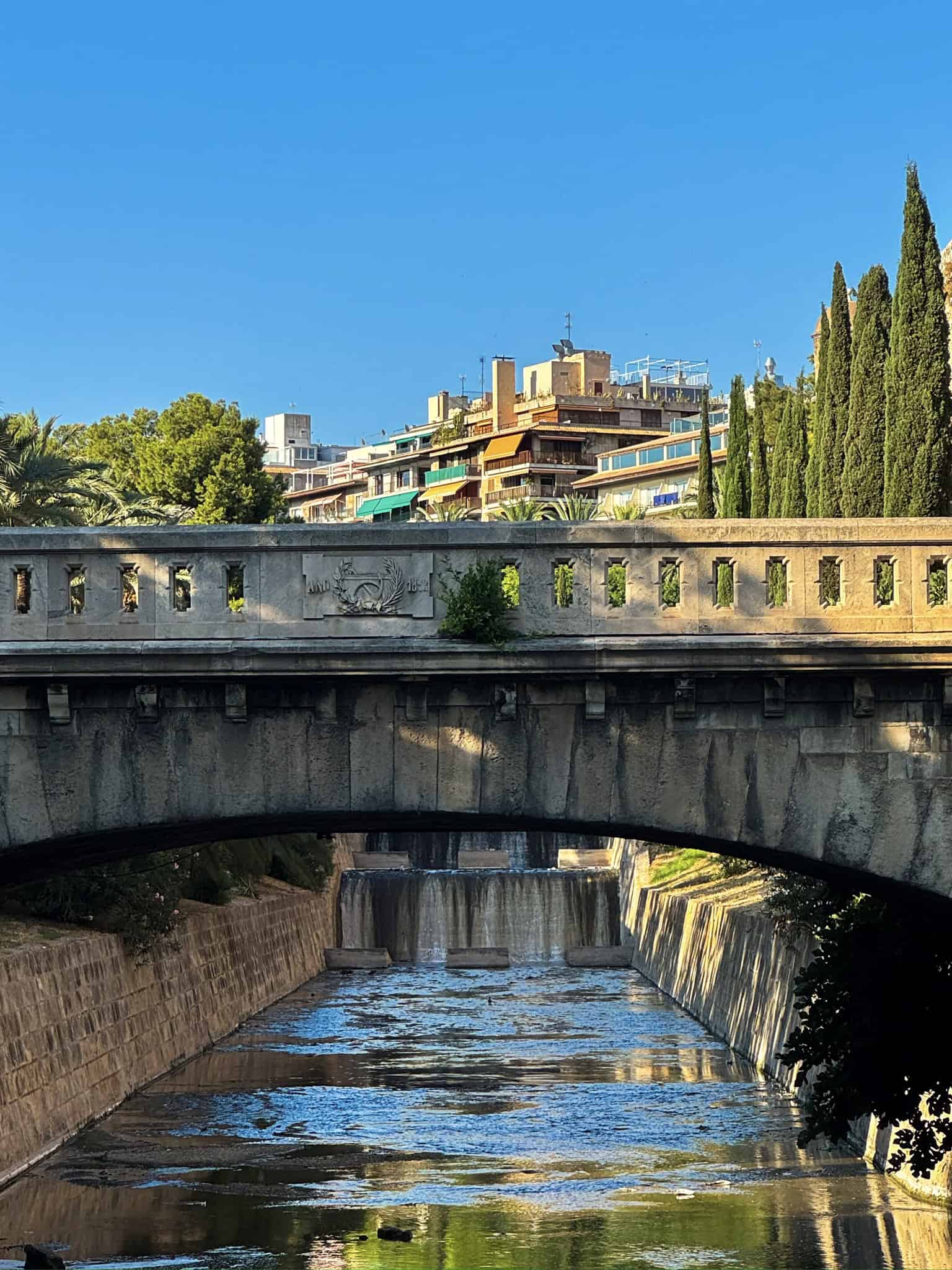 Picturesque view of a city canal with a stone bridge, surrounded by lush greenery and residential buildings against a clear blue sky in a sunny day.