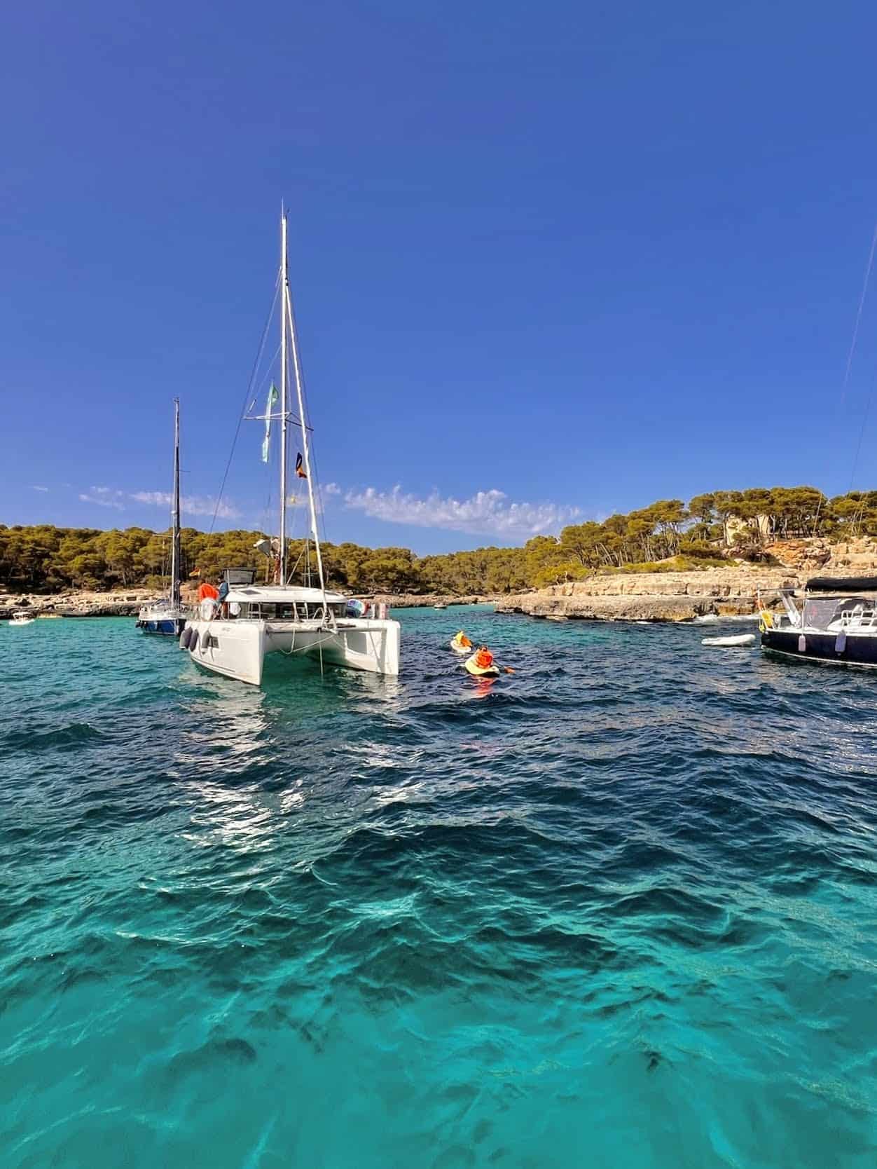 Sailboat anchored in clear turquoise waters near a rocky coastline with lush trees under a bright blue sky.