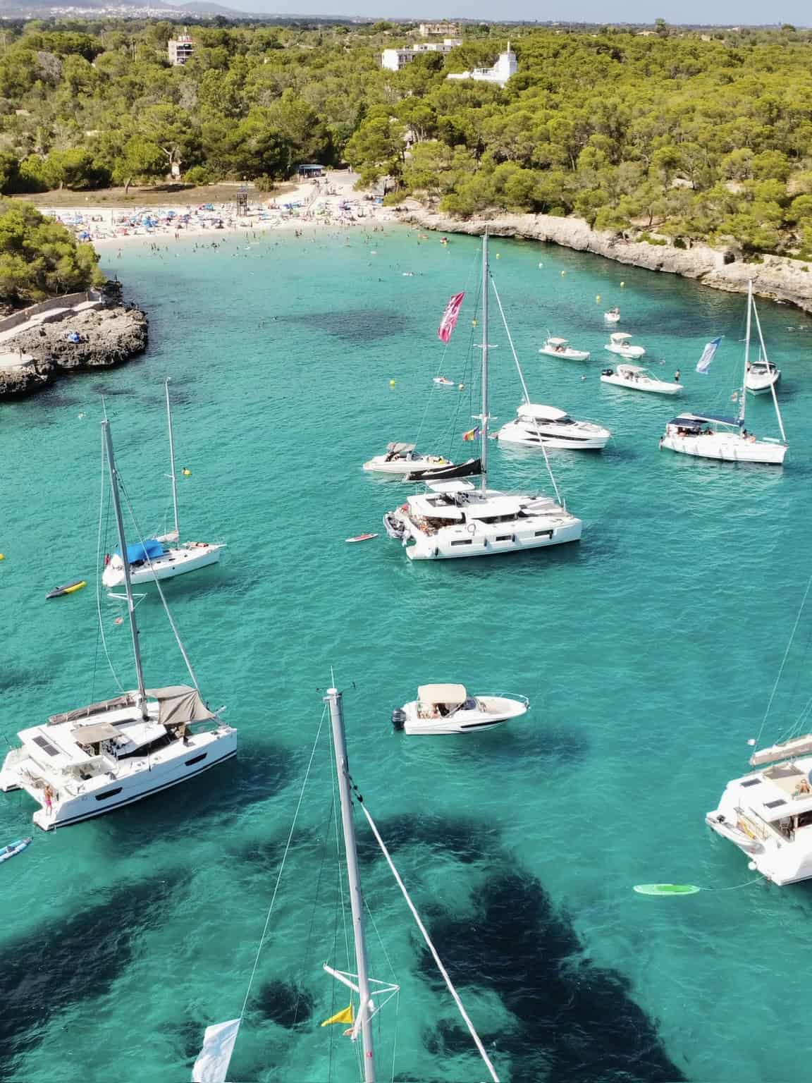 Luxury sailboats and yachts anchored in clear turquoise waters near a lush green coastal area, ideal for sailing holidays and maritime recreation in a scenic seaside destination.