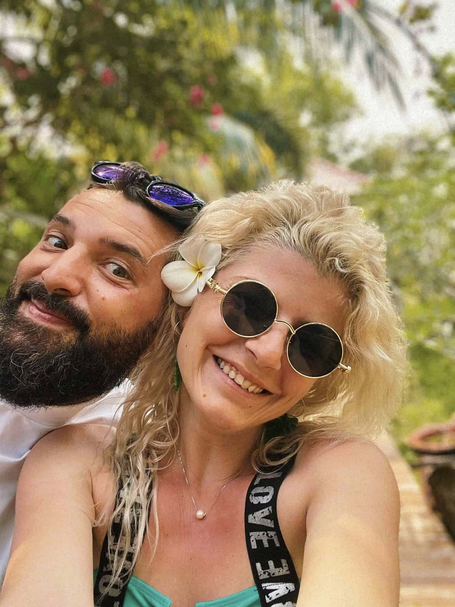 Smiling couple enjoying outdoor moment, woman with sunglasses and flower, man with sunglasses, lush greenery background.