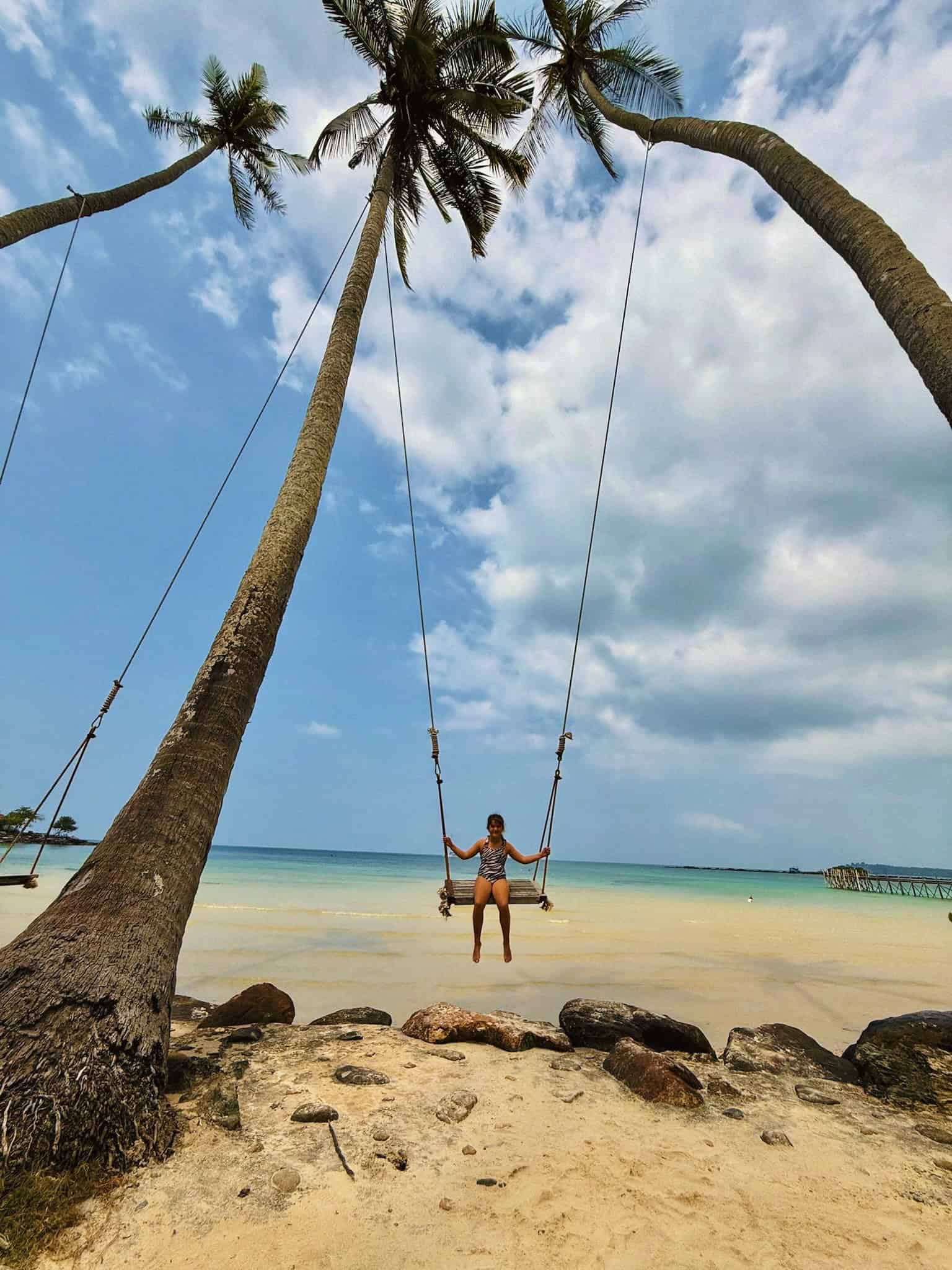 Swing on tropical palm trees by the beach with turquoise water and blue sky, perfect for relaxing seaside holiday, featuring a woman enjoying a swing among lush greenery.