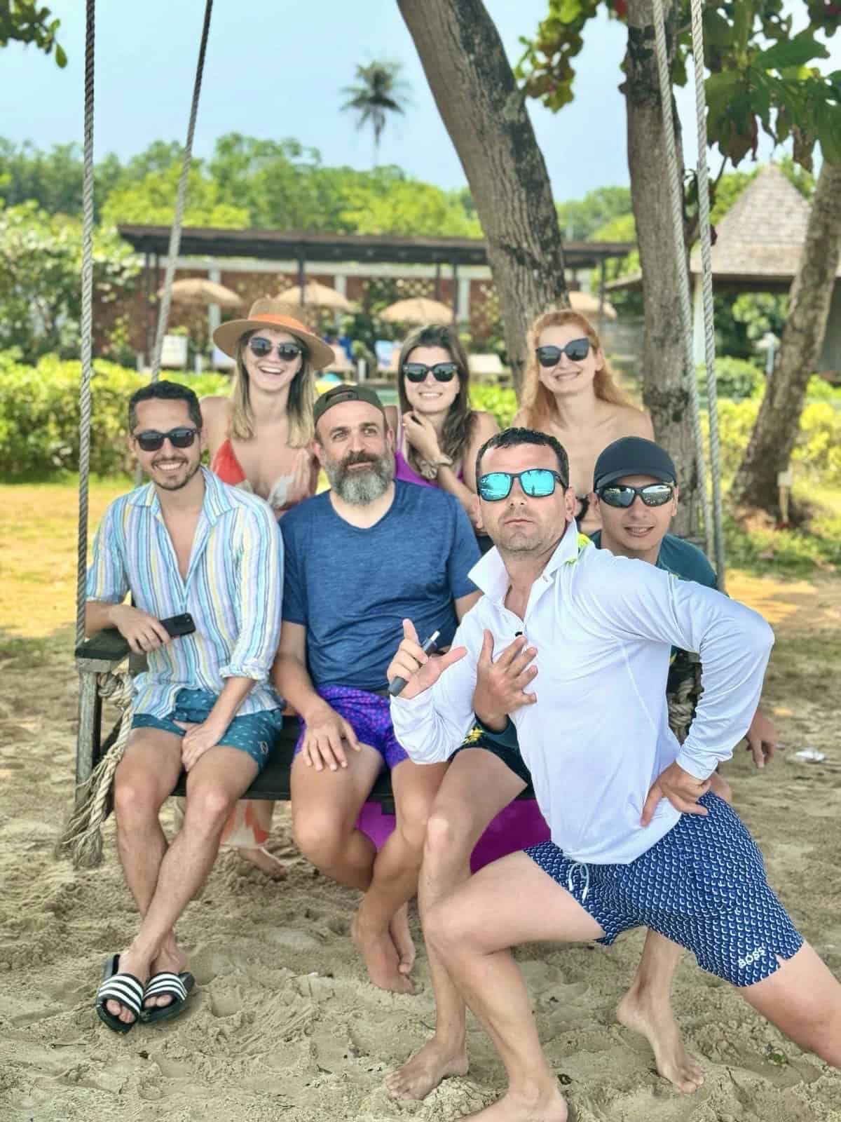 People enjoying a sunny day at the beach, sitting on a wooden swing surrounded by lush green trees and tropical scenery, dressed in colourful summer clothes and sunglasses for a fun, relaxing outdoor experience.