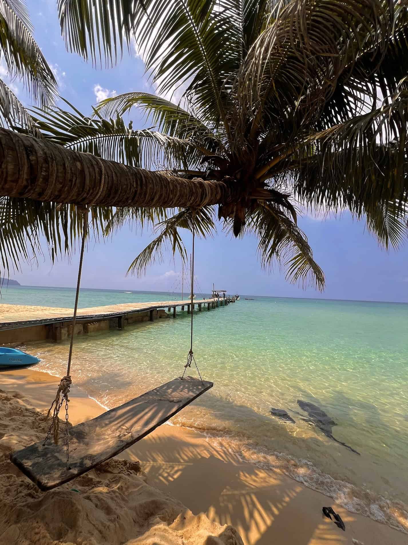 A tropical beach scene featuring a palm tree with a swing hanging from it, overlooking clear turquoise waters and a wooden pier extending into the ocean under a blue sky with some clouds.