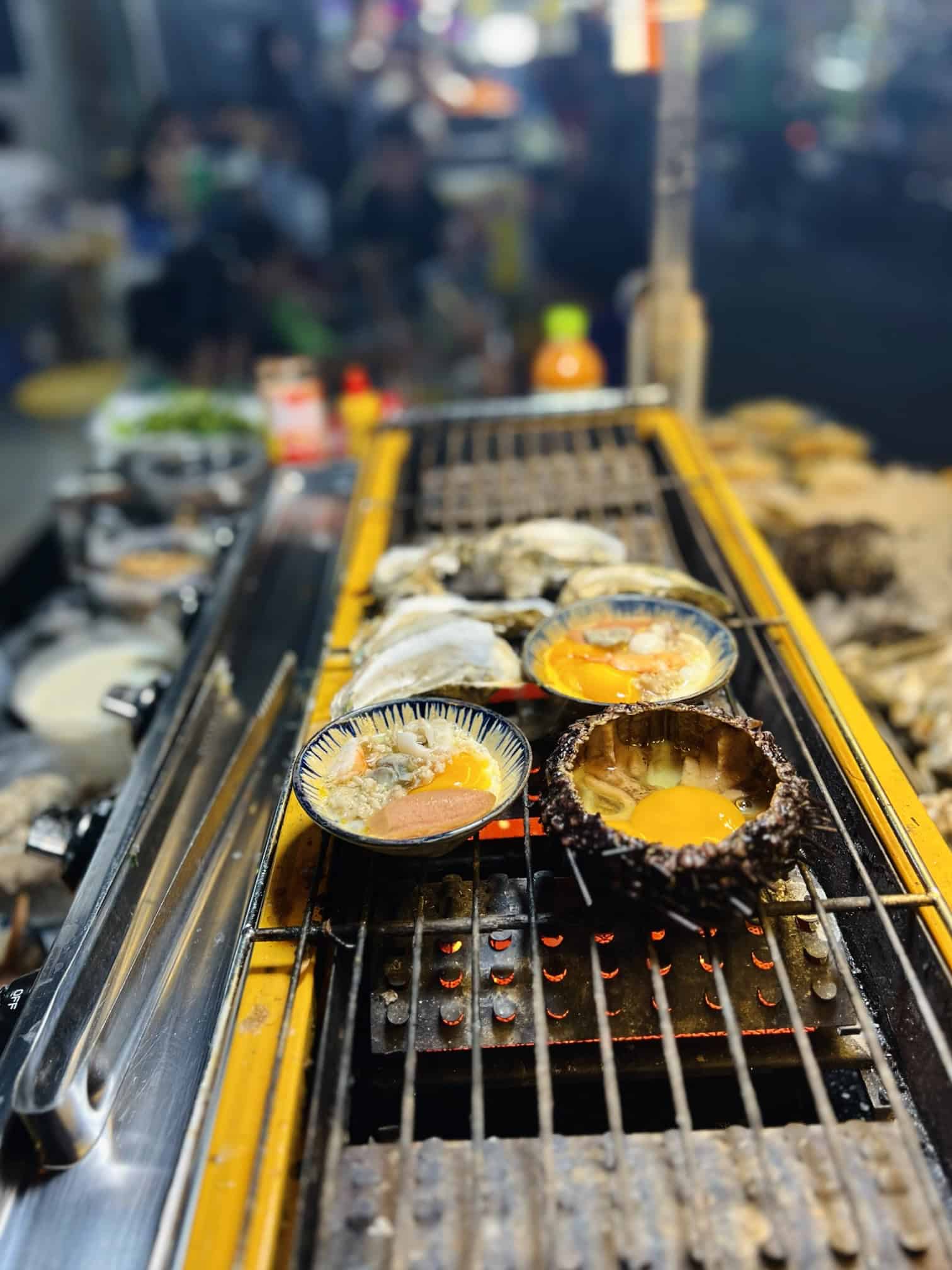 Fresh seafood grilling at an outdoor market, featuring oysters and eggs on a barbecue grill with vibrant market background, showcasing delicious and authentic seaside cuisine.