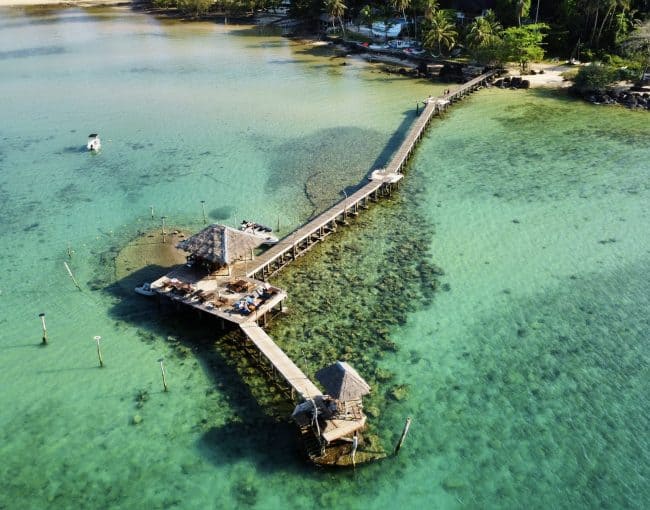 Boating pier extending into clear turquoise waters with a thatched-roof gazebo and surrounding tropical shoreline, perfect for relaxing seaside holidays and water activities.
