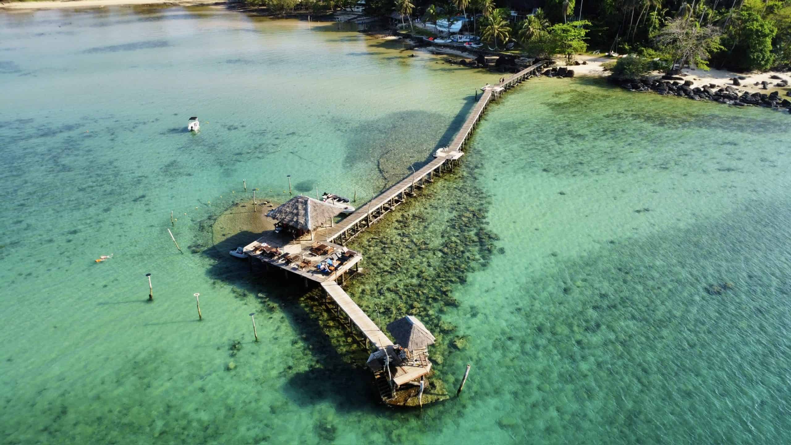 Boating pier extending into clear turquoise waters with a thatched-roof gazebo and surrounding tropical shoreline, perfect for relaxing seaside holidays and water activities.