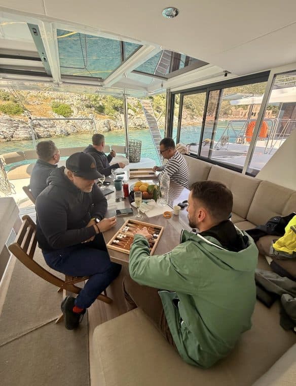 Relaxing boat interior with friends enjoying a casual game of backgammon by the water. Large glass windows showcase a scenic view of the sea and rocky shoreline, creating a perfect setting for leisure and socialising onboard.