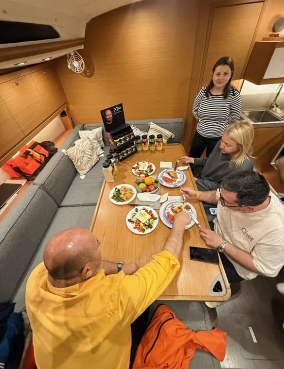 Coffee table with fresh fruit, salads, and beverages on a yacht interior during a social gathering, with people enjoying food and drinks, highlighting luxury and leisure.