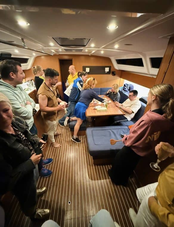 Cozy yacht interior during social gathering with people around dining table, warm lighting, wooden accents, casual atmosphere, socialising and celebration on luxury vessel.