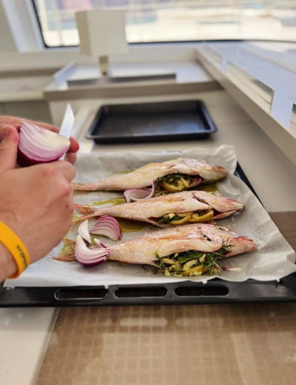 Fresh fish being prepared with herbs and onions for baking or grilling, displayed on parchment paper on a baking tray in a modern kitchen setting.