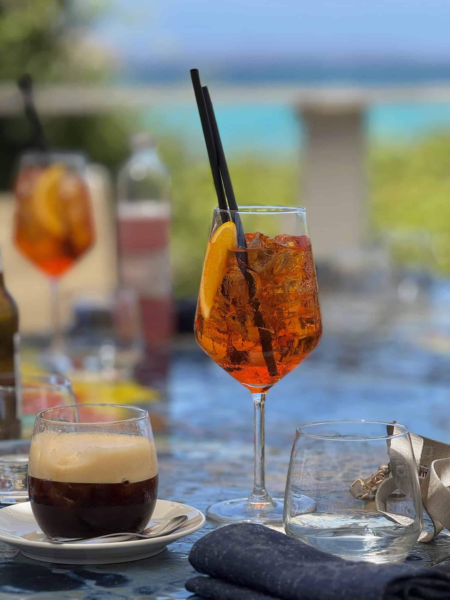 A vibrant cocktail with orange slices and black straws on a table outdoors, with a blurred lakeside background and other drinks, perfect for summer refreshment and relaxation.