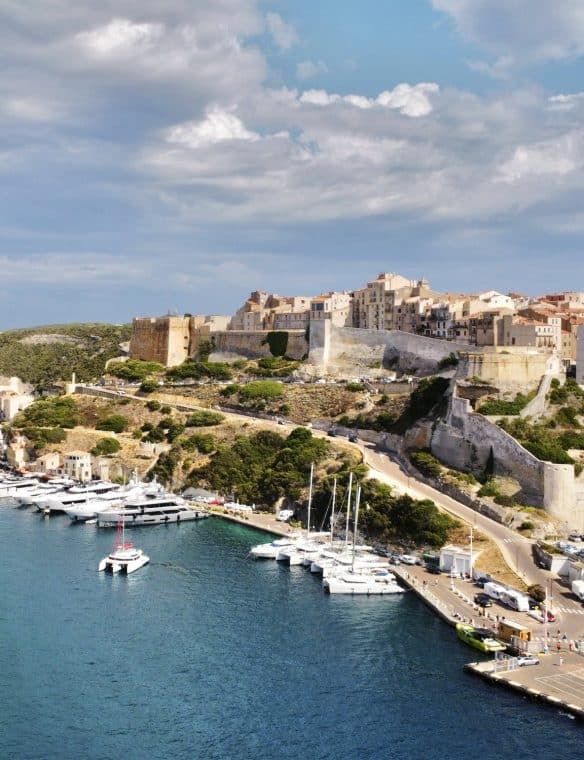 Picturesque view of a historic coastal city with an ancient fortress and colourful buildings overlooking a harbour filled with yachts and boats under a partly cloudy sky.