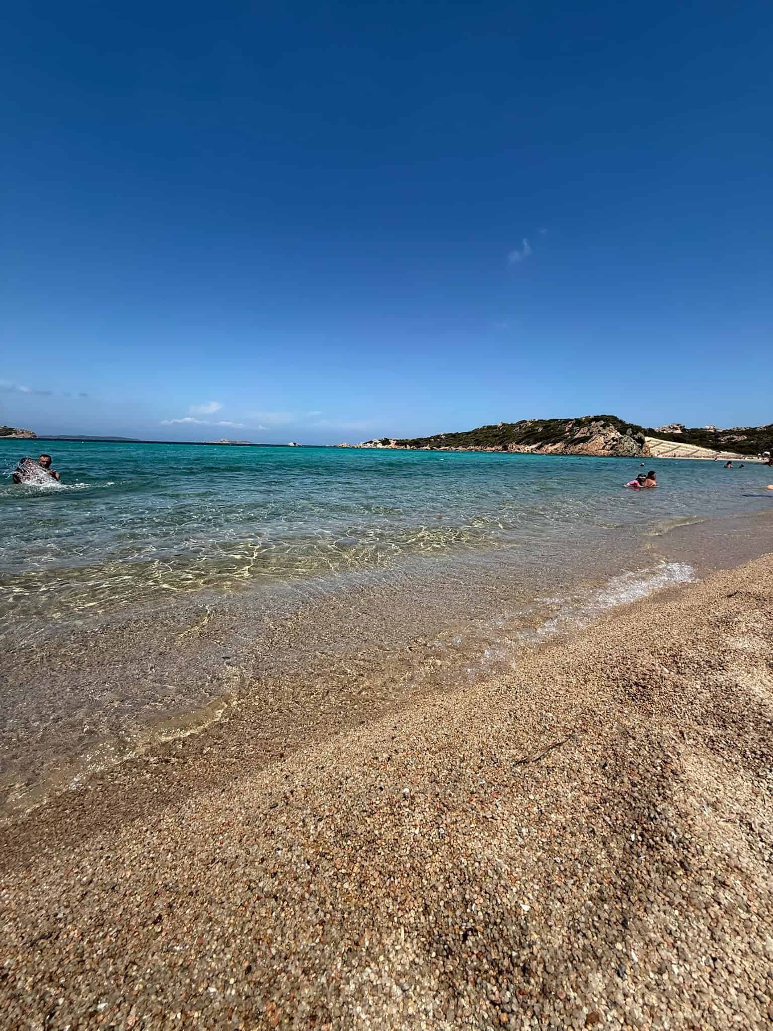Vibrant beach scene with clear turquoise waters, sandy shore with small pebbles, blue sky, and rocky islands in the distance; ideal for seaside holiday memories.