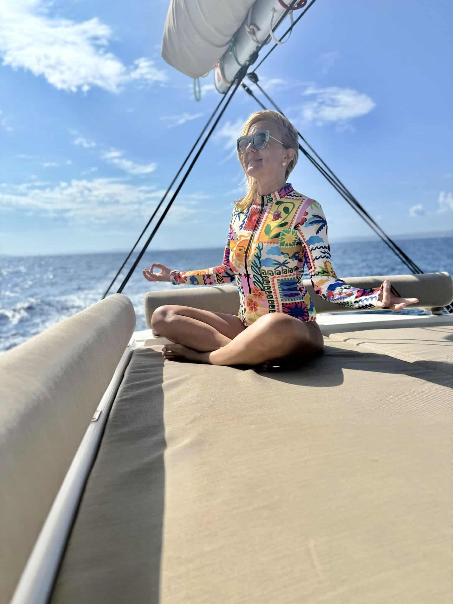 Relaxing woman doing yoga on a yacht in the open sea, wearing colourful tropical print clothing, with blue sky and ocean background, emphasising wellness, leisure, and adventure.