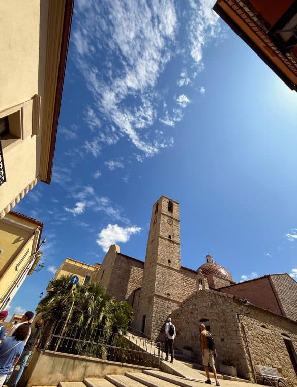 Ancient stone church tower with a clock, surrounded by historic buildings and lush greenery, under a bright blue sky with scattered clouds, in a popular European tourist destination.