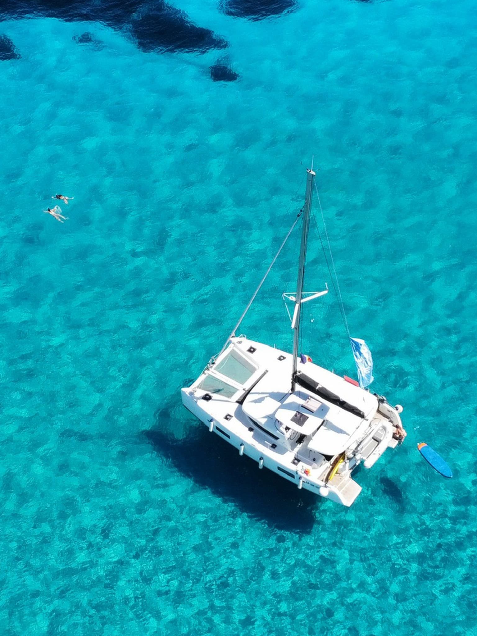 Aerial view of a luxury white sailing yacht anchored in clear turquoise waters, with two people swimming nearby and a colourful paddleboard floating beside the boat.