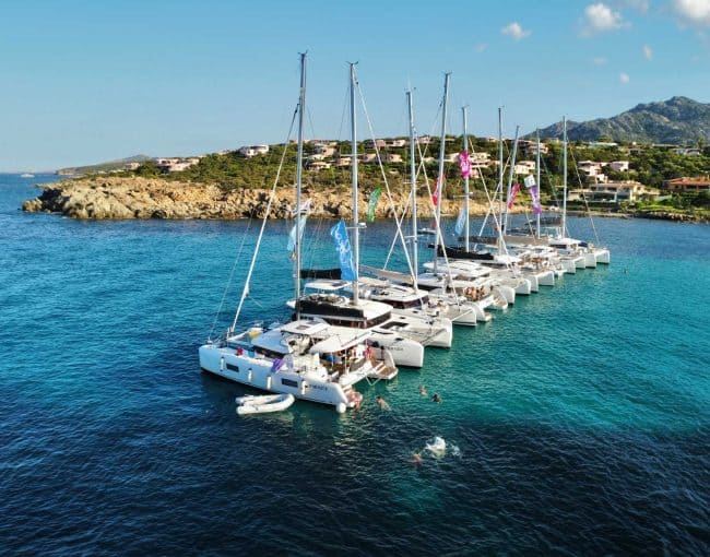 Luxury sailboats docked in a scenic bay with clear turquoise water and hillside residential area in the background.