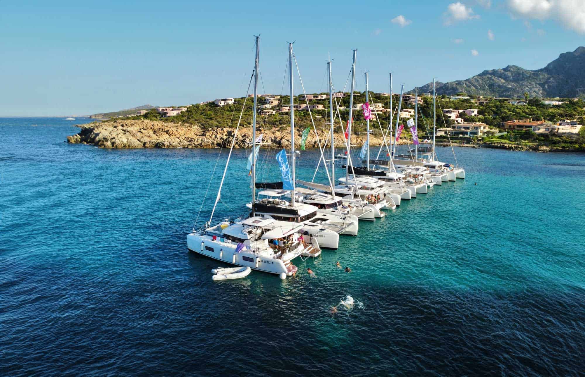 Luxury sailboats docked in a scenic bay with clear turquoise water and hillside residential area in the background.