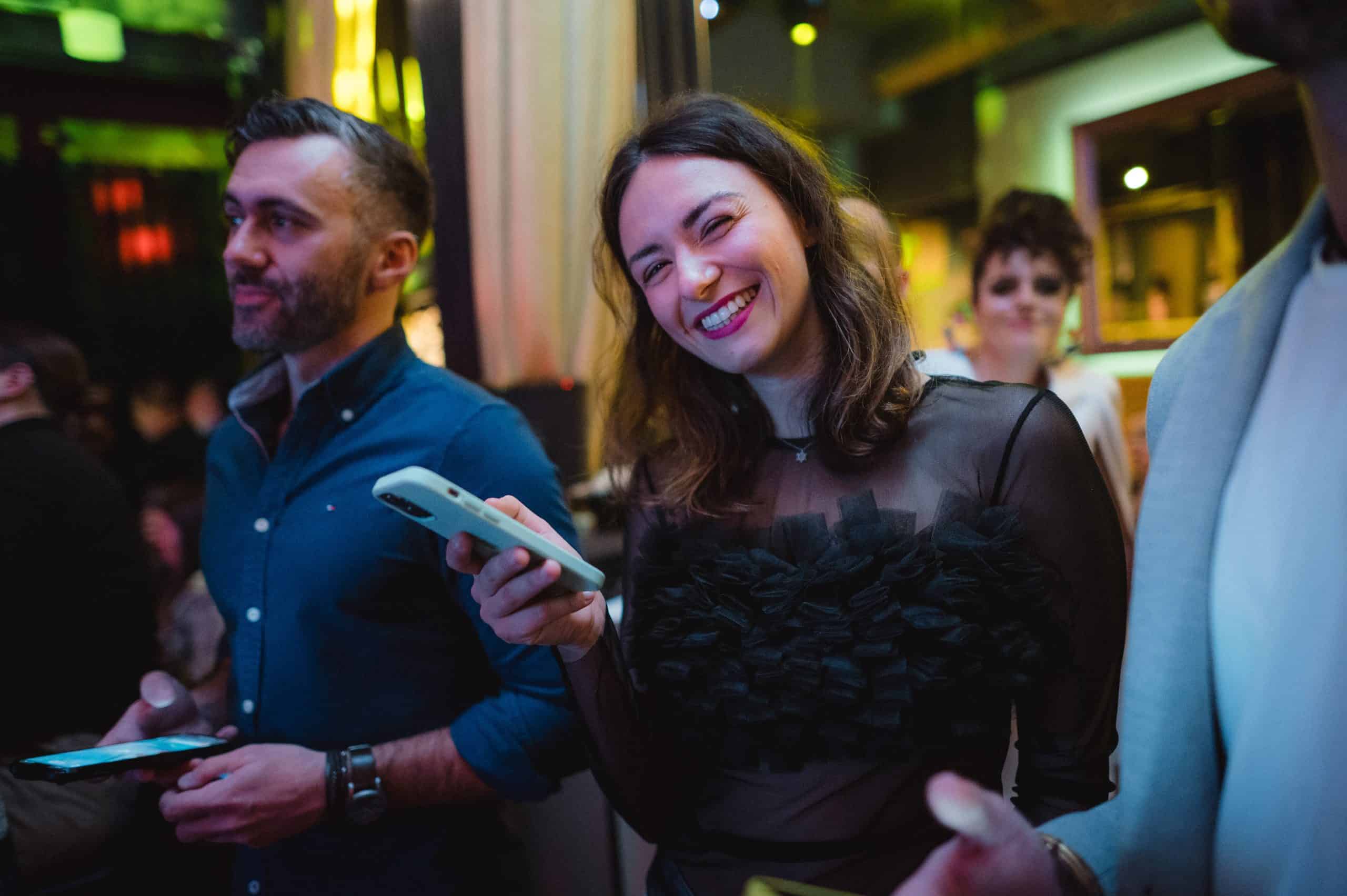 People enjoying socialising at a lively event, with a woman smiling holding a smartphone, highlighting vibrant nightlife, entertainment, and community engagement.