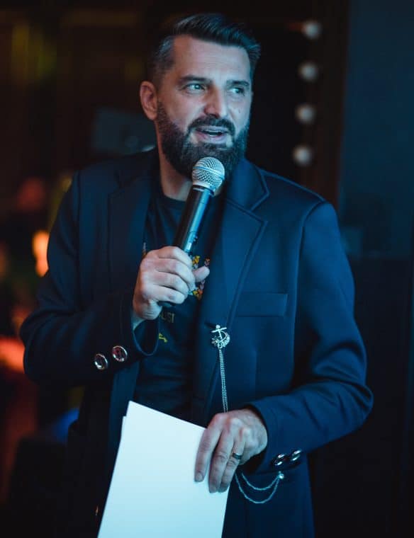 Professional man speaking at an event, holding a microphone and a white sheet of paper, dressed in a black blazer with a unique chain accessory, in a modern venue.