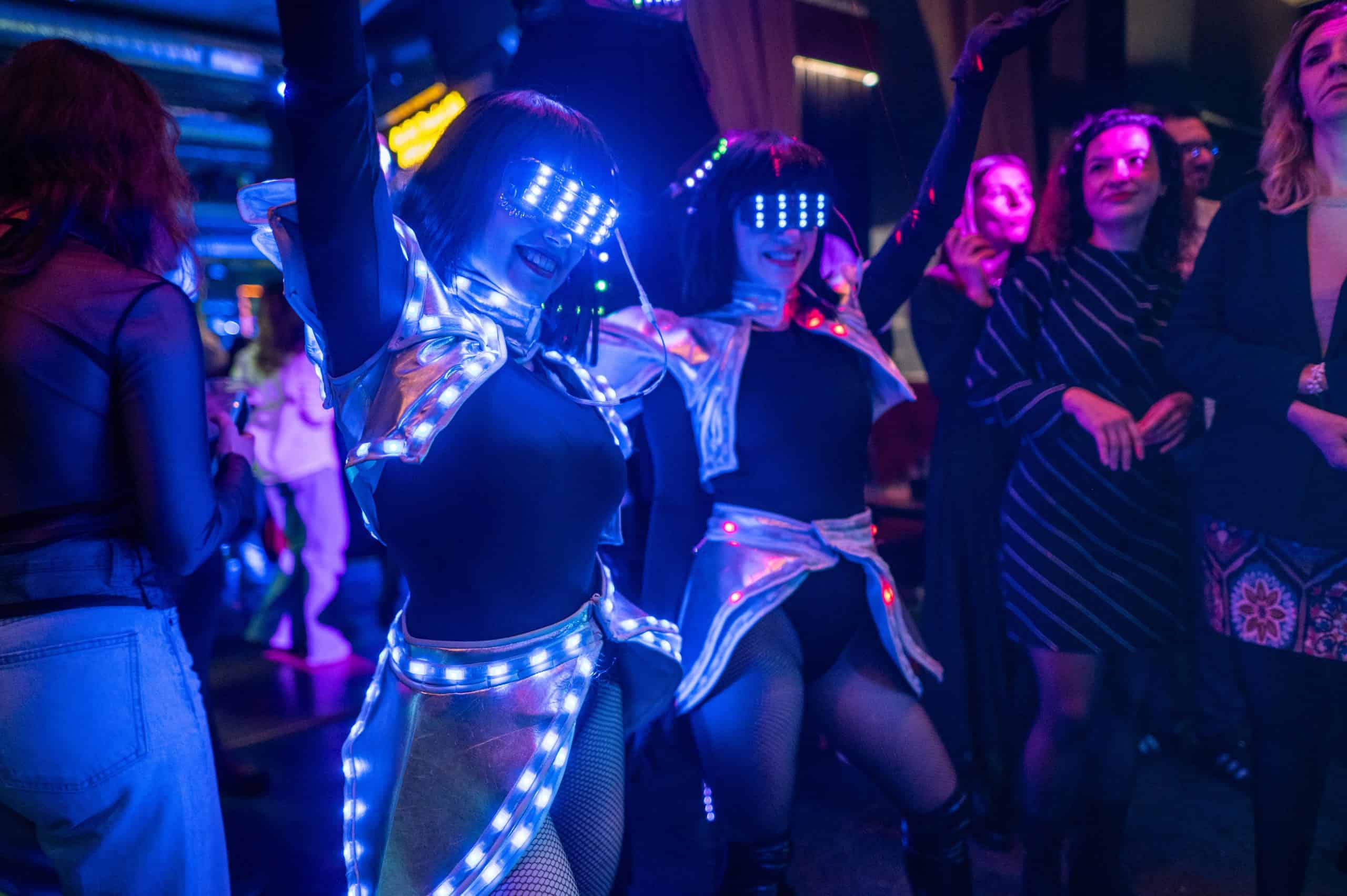 Vibrant nightlife scene with women wearing LED illuminated futuristic costumes at a dance party, showcasing colourful lighting and energetic atmosphere.