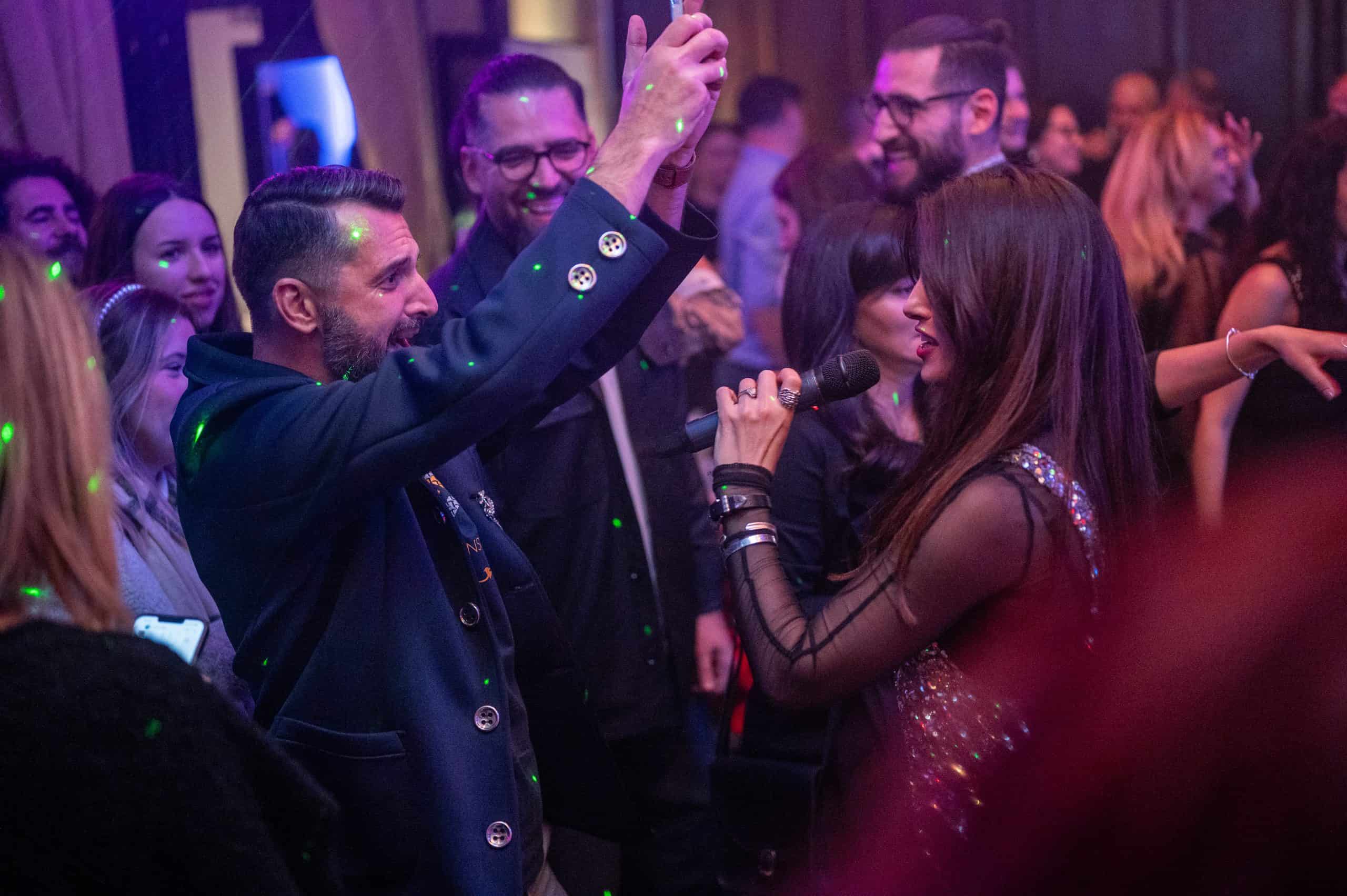 Dancing crowd at a lively party, with a woman singing into a microphone and a man raising his hands, surrounded by partygoers in a vibrant, colourful nightclub setting.