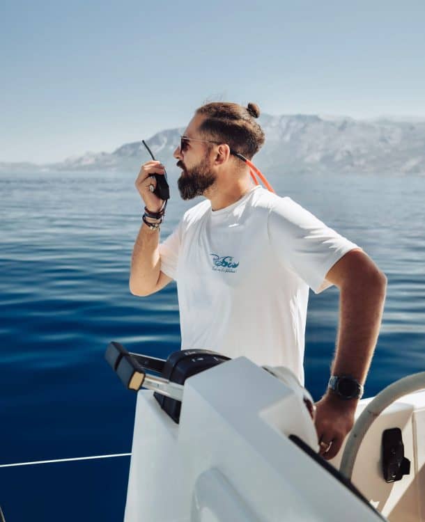 An experienced man on a boat using a radio communication device, with a scenic ocean and mountain background, demonstrating maritime communication skills.